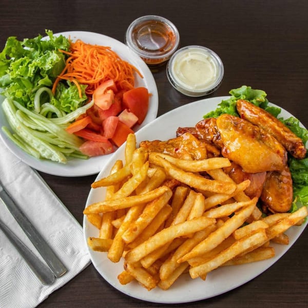 Plates of golden french fries, glazed chicken wings, and a fresh salad with dressing on a dark table.