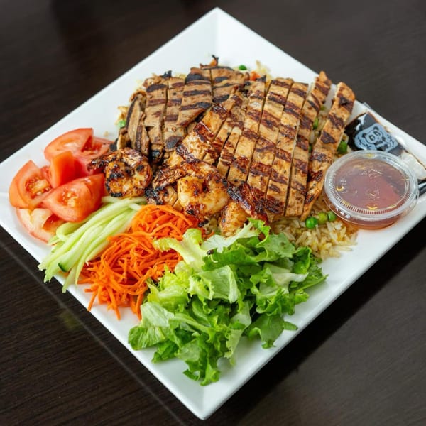 Grilled chicken, beef, and shrimp with rice, fresh vegetables, and sauce on a white plate.