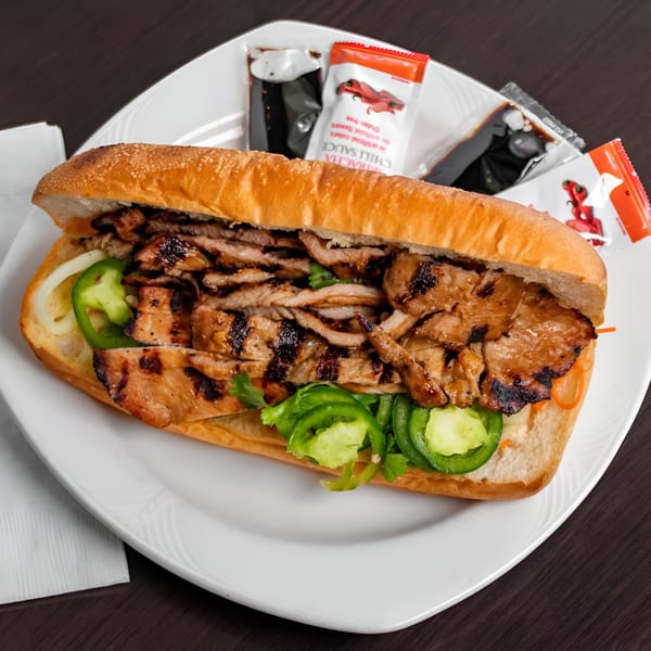 Grilled pork banh mi sandwich with jalapeños, cilantro, and sauces on a white plate.