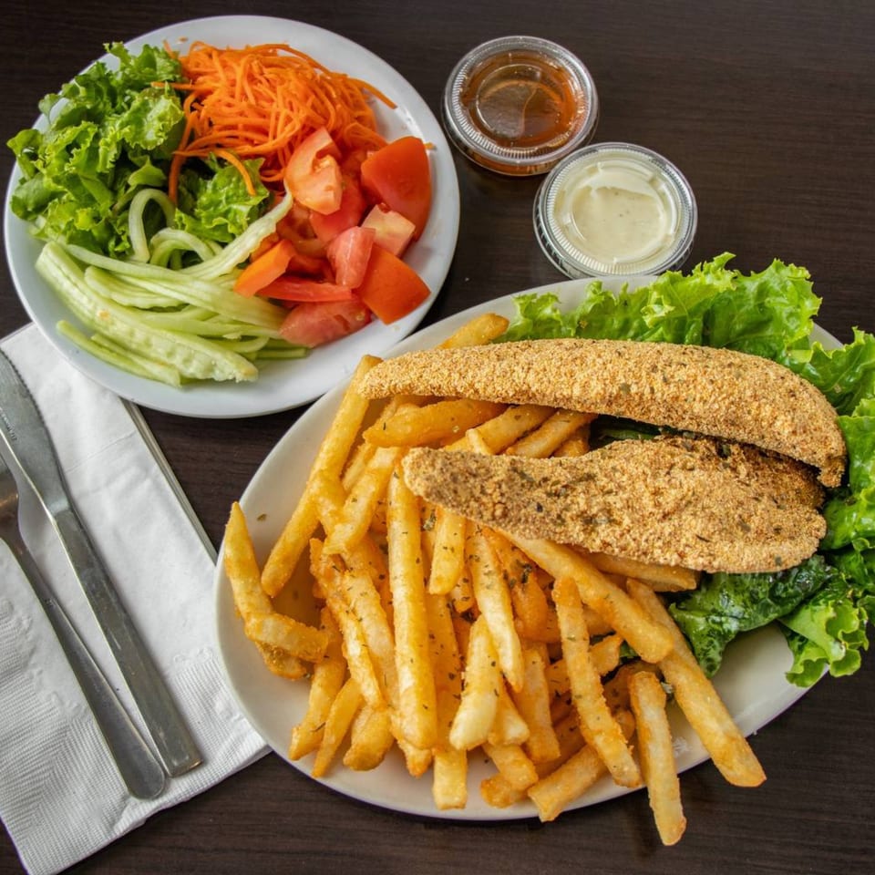 Fried fish fillets with french fries and a side salad with dressing cups on a dark table.
